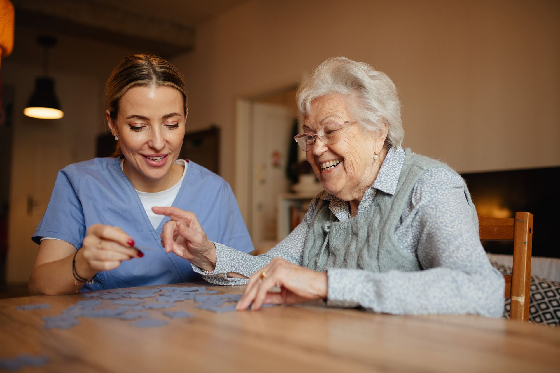 Caregiver in blue scrubs assists an older person playing a game at a wooden table. Both smile.