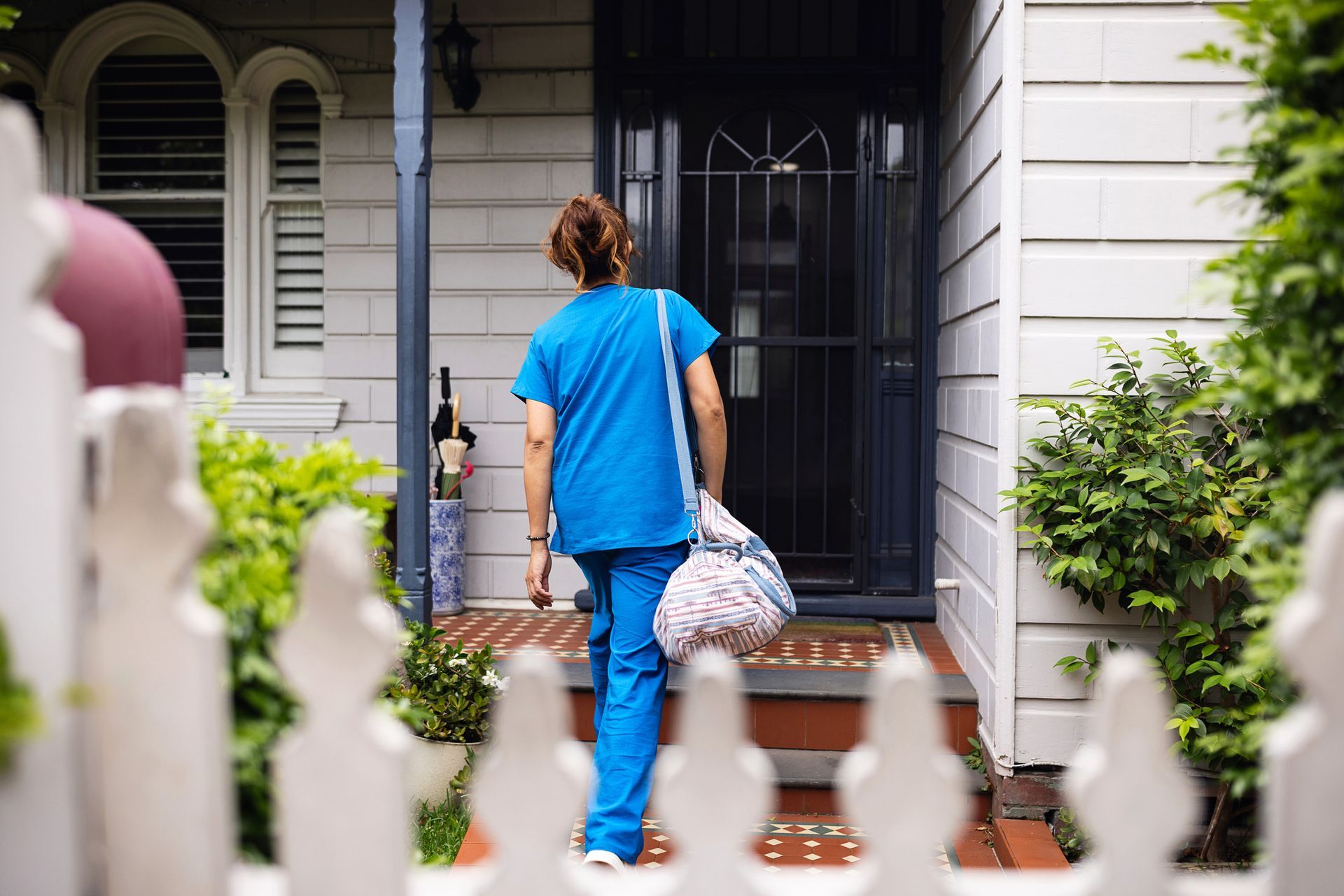 Person in blue scrubs enters a house, carrying a bag. A white picket fence is in the foreground.