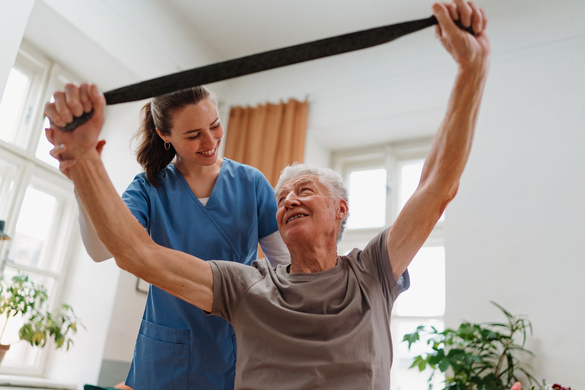 Physical therapist assisting a smiling elderly man with stretching using a resistance band indoors.
