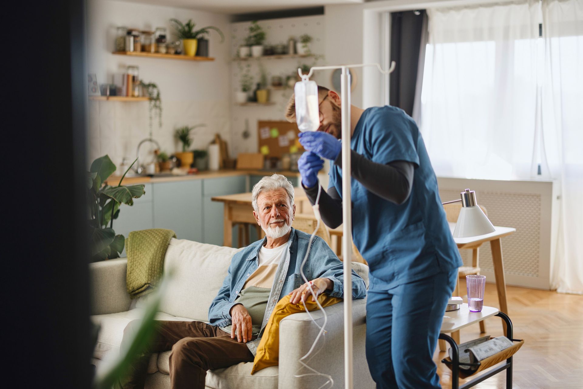 Man receiving IV treatment at home; nurse in blue scrubs adjusts the bag.
