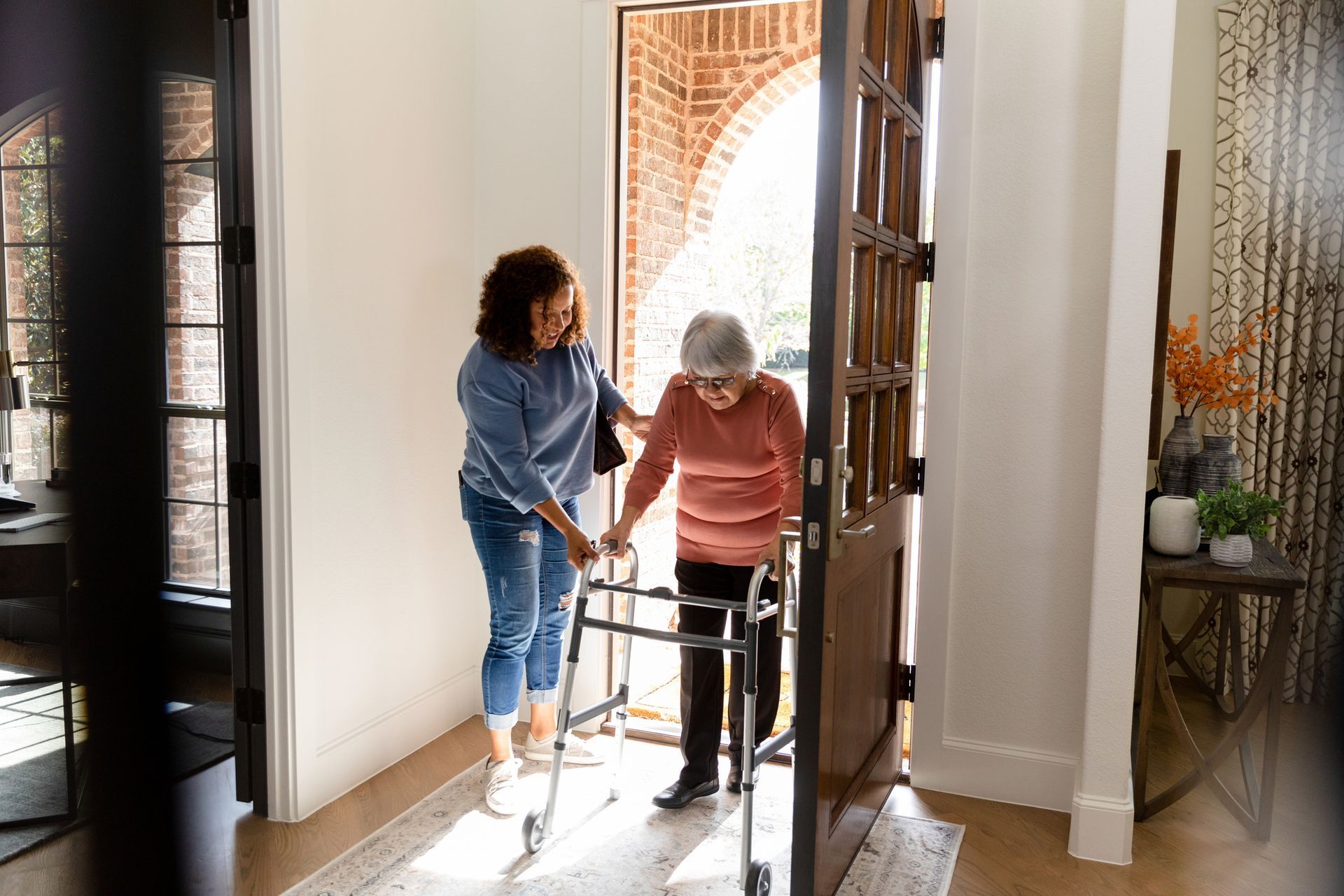 Woman helping elderly woman with walker through doorway.