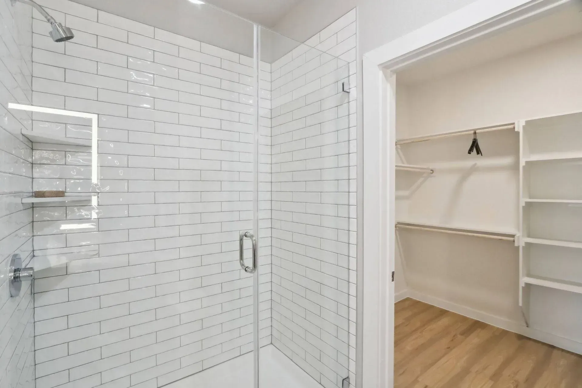 White-tiled shower with glass door next to a walk-in closet with shelves and wood-look flooring.