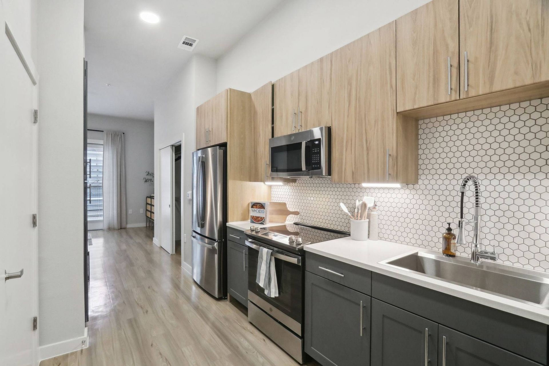 Modern apartment kitchen with stainless steel appliances, wood upper cabinets, and a white hex tile backsplash.