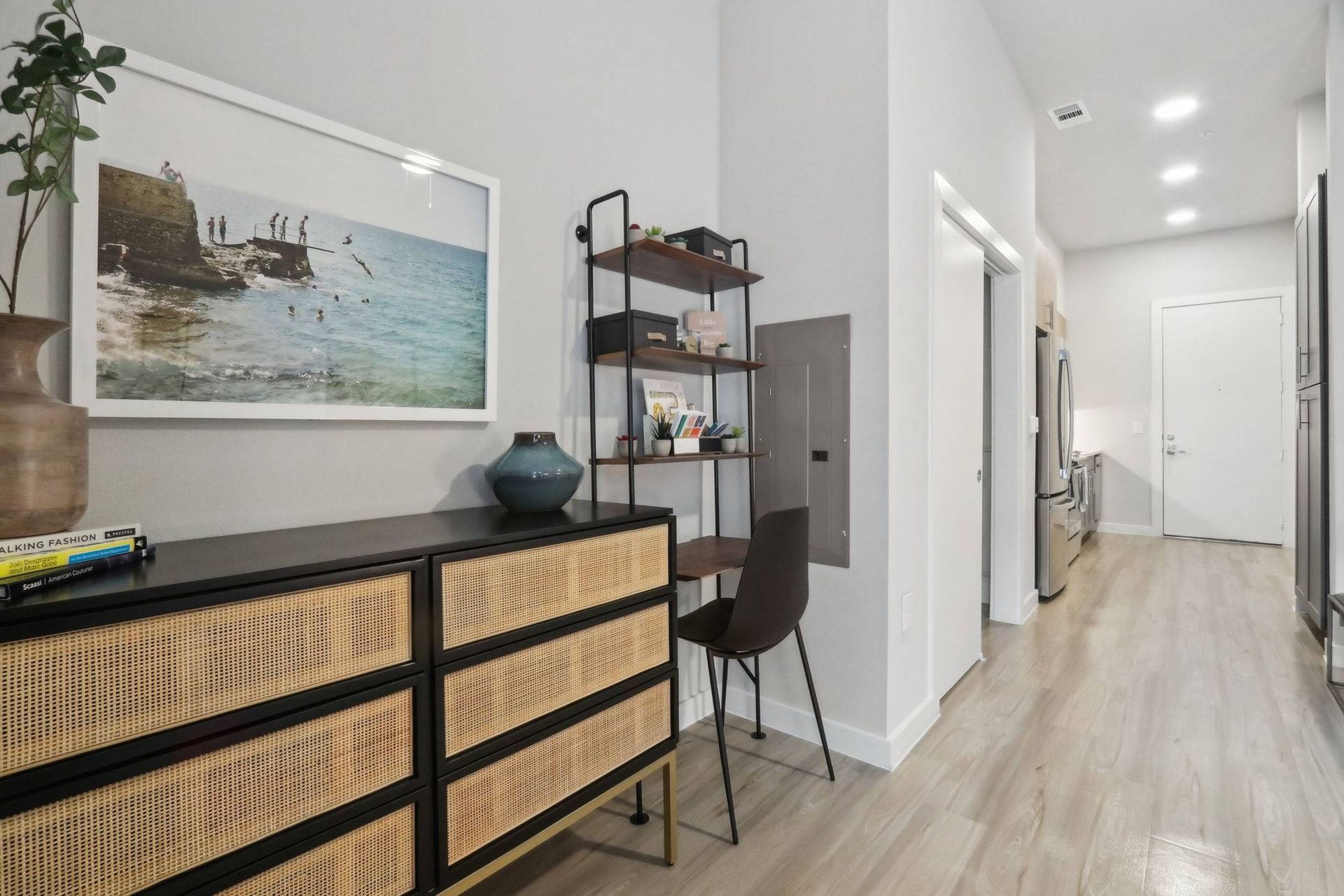 Modern apartment interior featuring a dresser, desk nook, shelving unit, and open kitchen at the end.