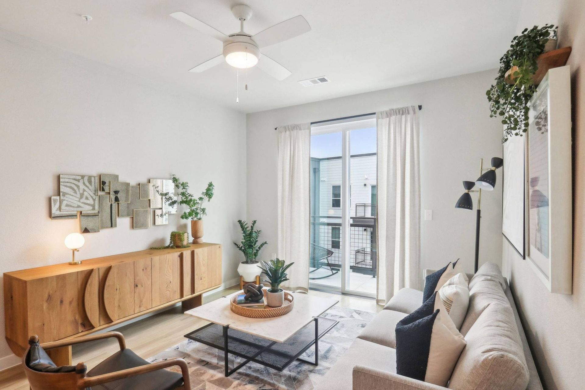 Living room in an apartment with a beige sofa, coffee table, plants, and a sliding glass balcony door.