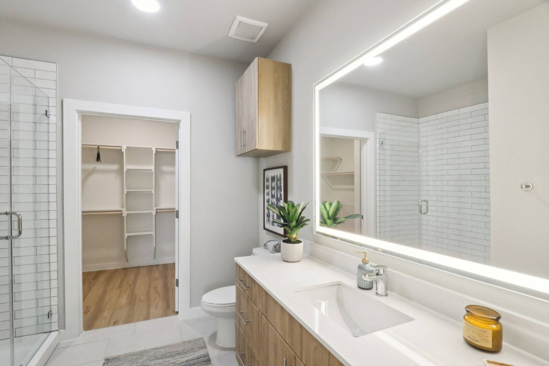 Bright bathroom with a double vanity, backlit mirror, glass shower, and a walk-in closet visible through an open doorway.