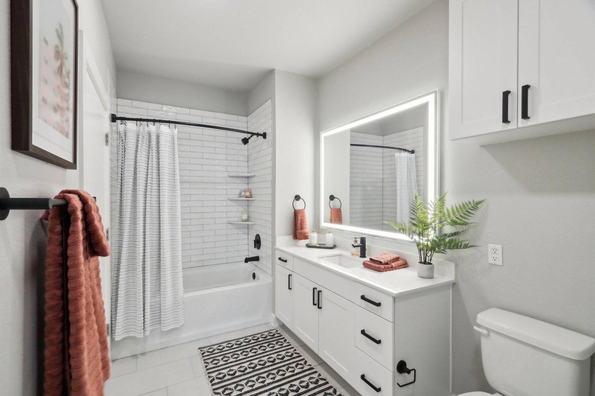 White bathroom with vanity, illuminated mirror, tub/shower, and plant.