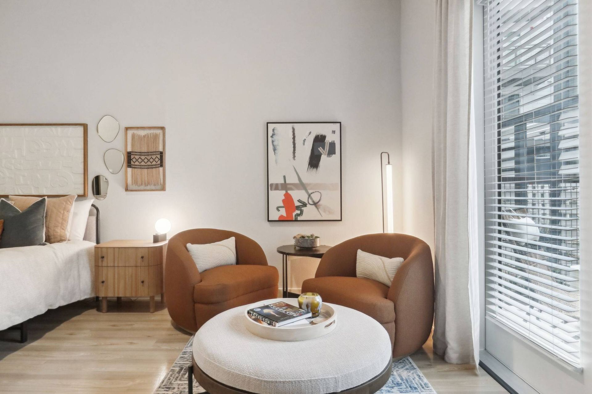 Living area with two brown curved chairs, a round ottoman, bed partially visible, and a large window with blinds.