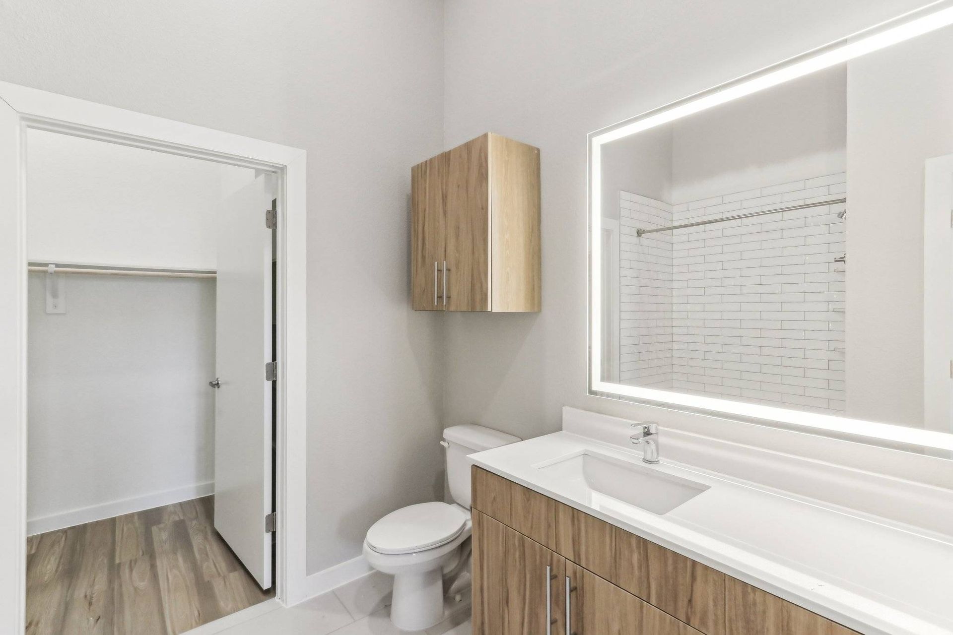 Bathroom in a modern apartment with a wood-toned vanity, backlit mirror, toilet, and an open closet with a hanging rod.