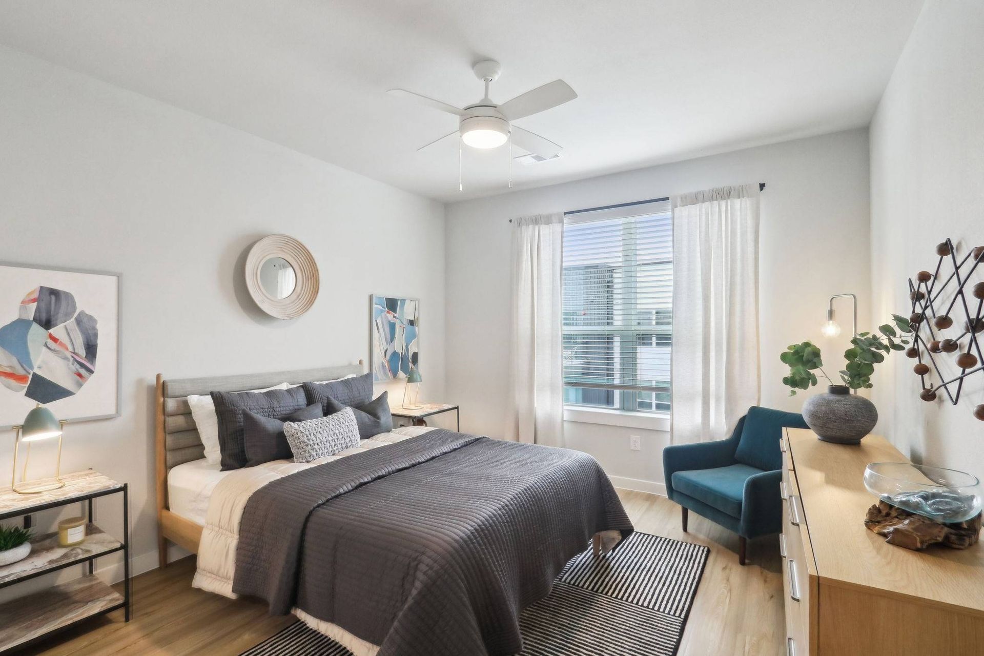 Furnished bedroom in an apartment with a bed, dresser, and window with sheer curtains.