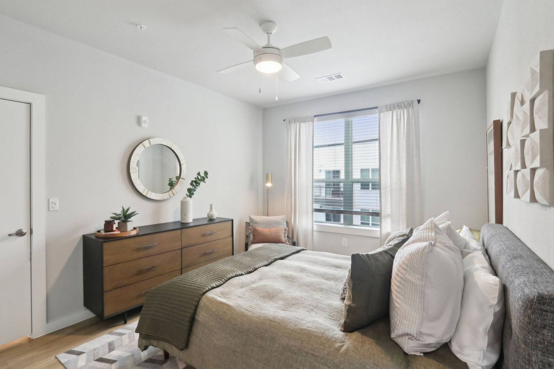 Bedroom in a modern apartment with a queen bed, dresser, round mirror, and a window with white curtains.