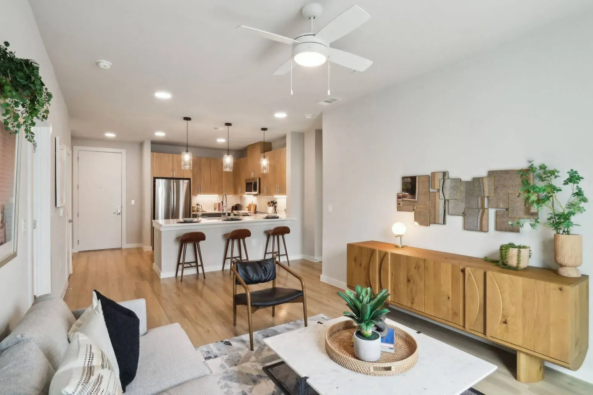 Open-concept apartment living room with light wood floors, kitchen with a breakfast bar, and a wooden credenza.