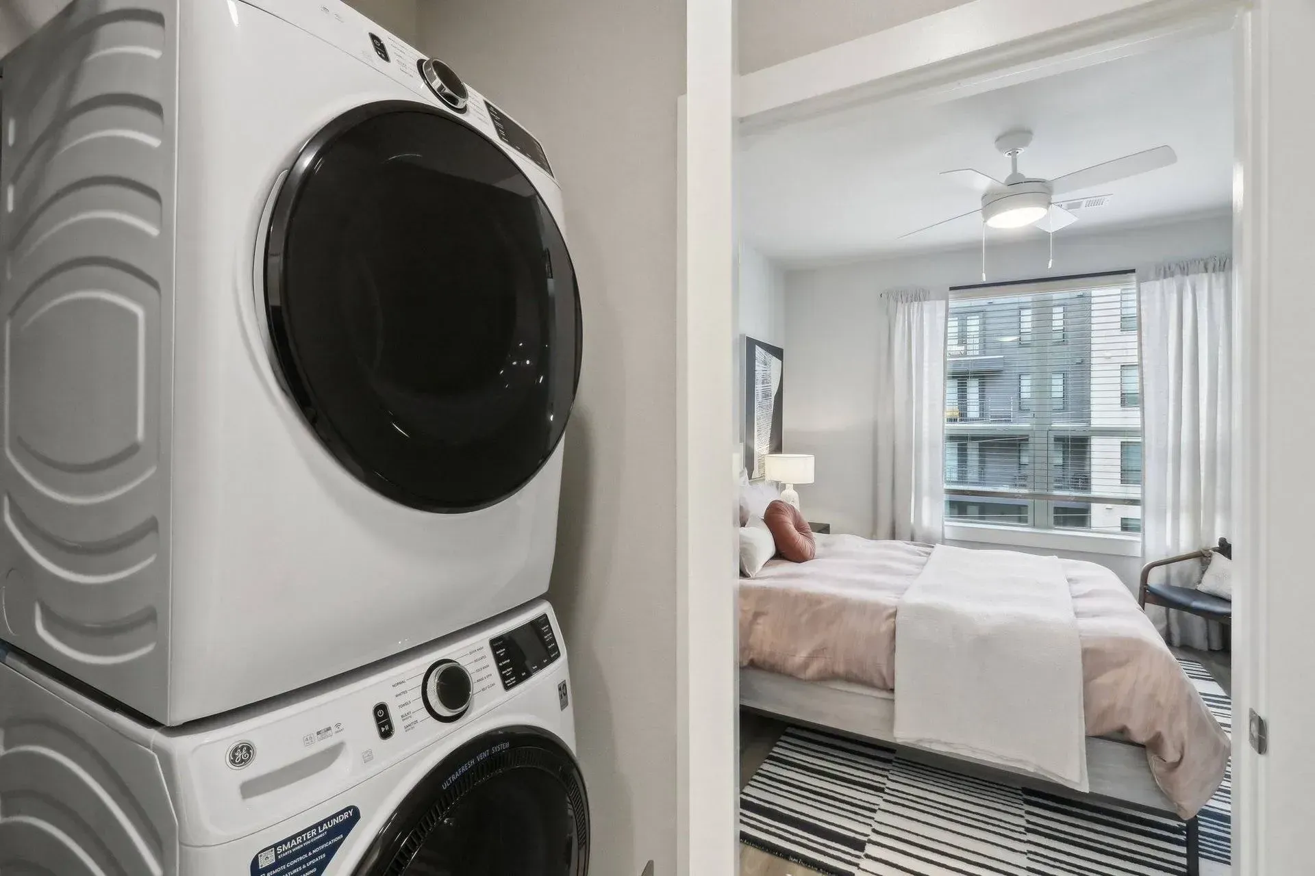Stacked white washer and dryer in a utility closet with a bedroom visible in the background.