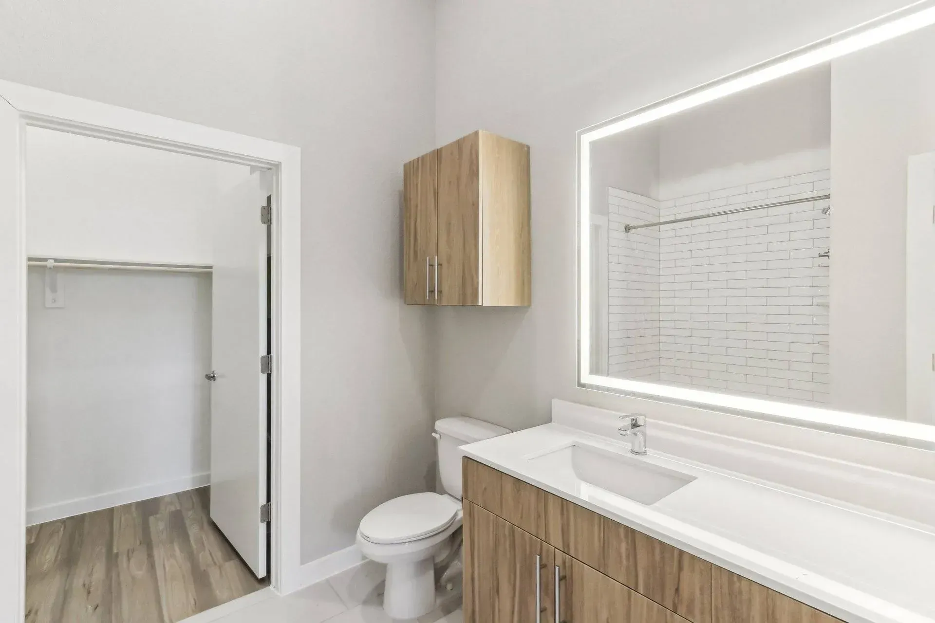 Bathroom with toilet, sink, mirror, and closet with wood-grain cabinets and light-colored walls.