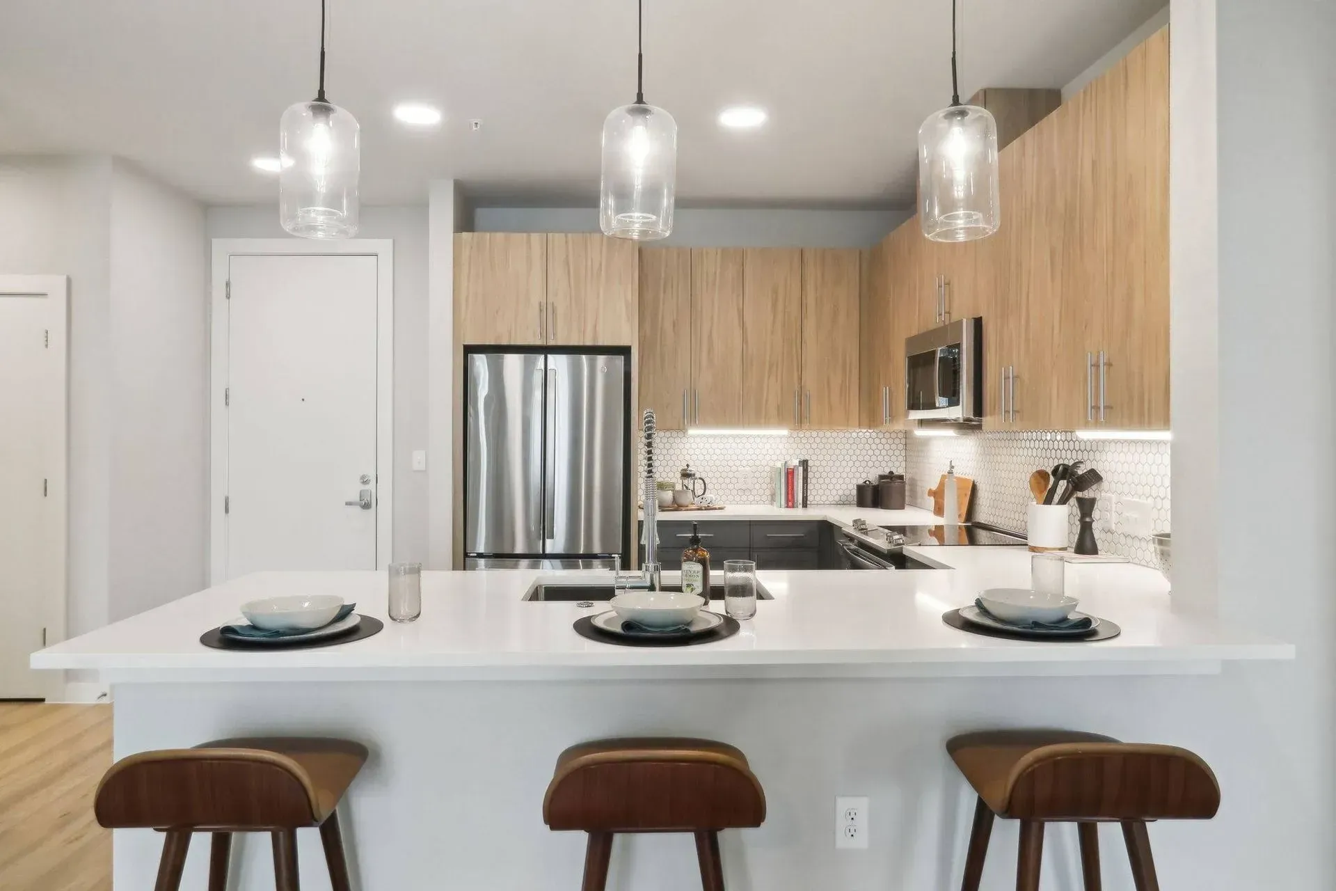 Modern kitchen with white countertops, wooden cabinets, and three bar stools.