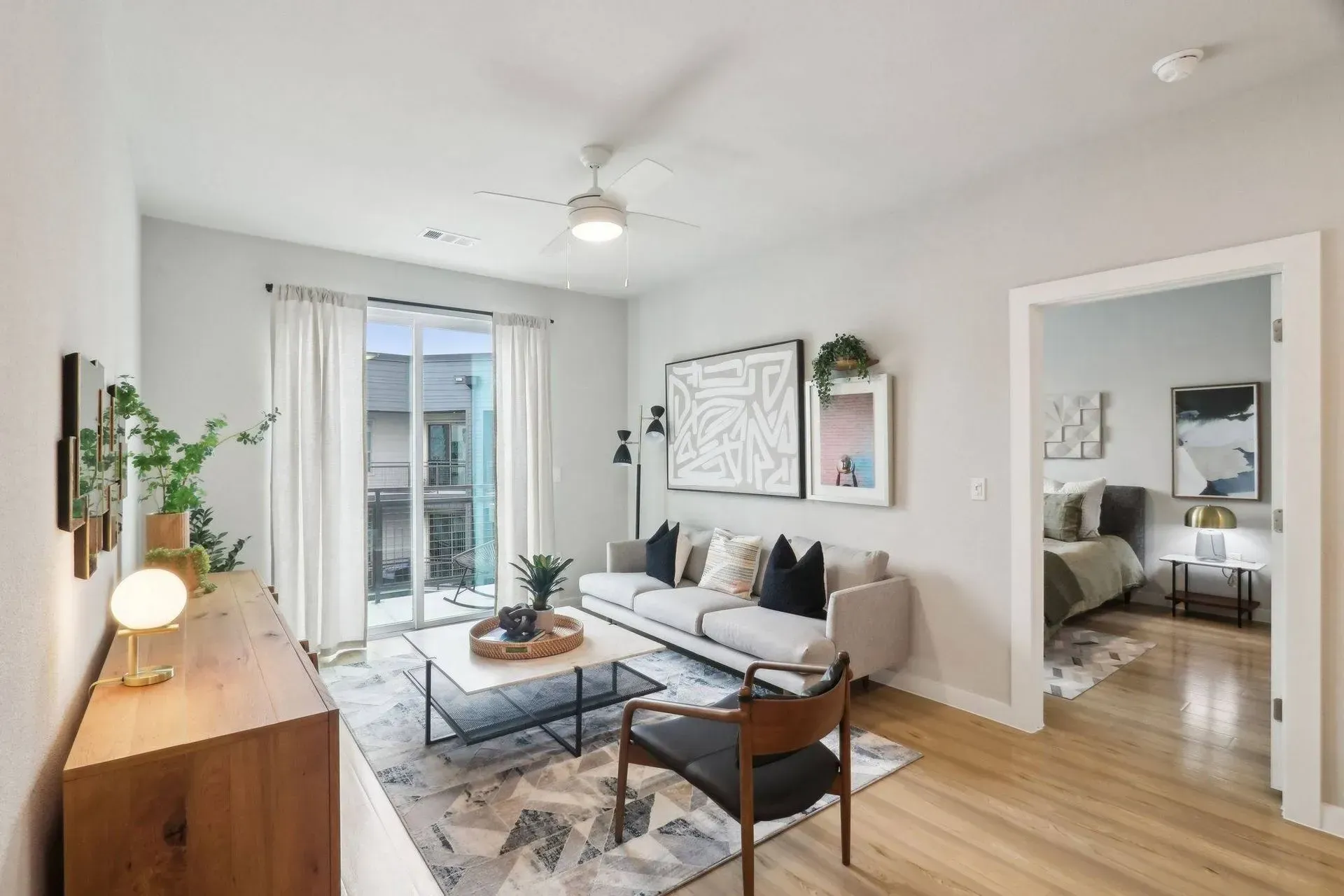 Modern living room with light wood floors, neutral walls, and access to a balcony; view into bedroom.