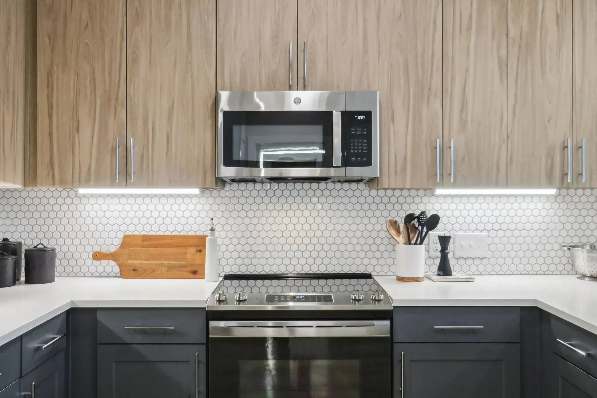 Modern kitchen with wood-tone upper cabinets, grey lower cabinets, white backsplash, and stainless steel appliances.