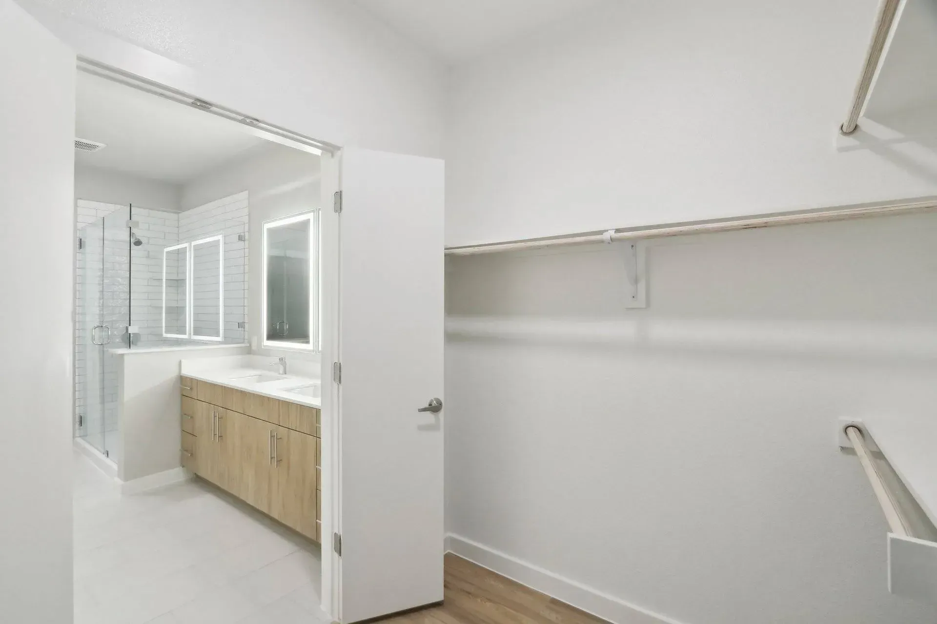 Walk-in closet with shelves and partial view of a bathroom with a vanity and shower. White walls and wood flooring.