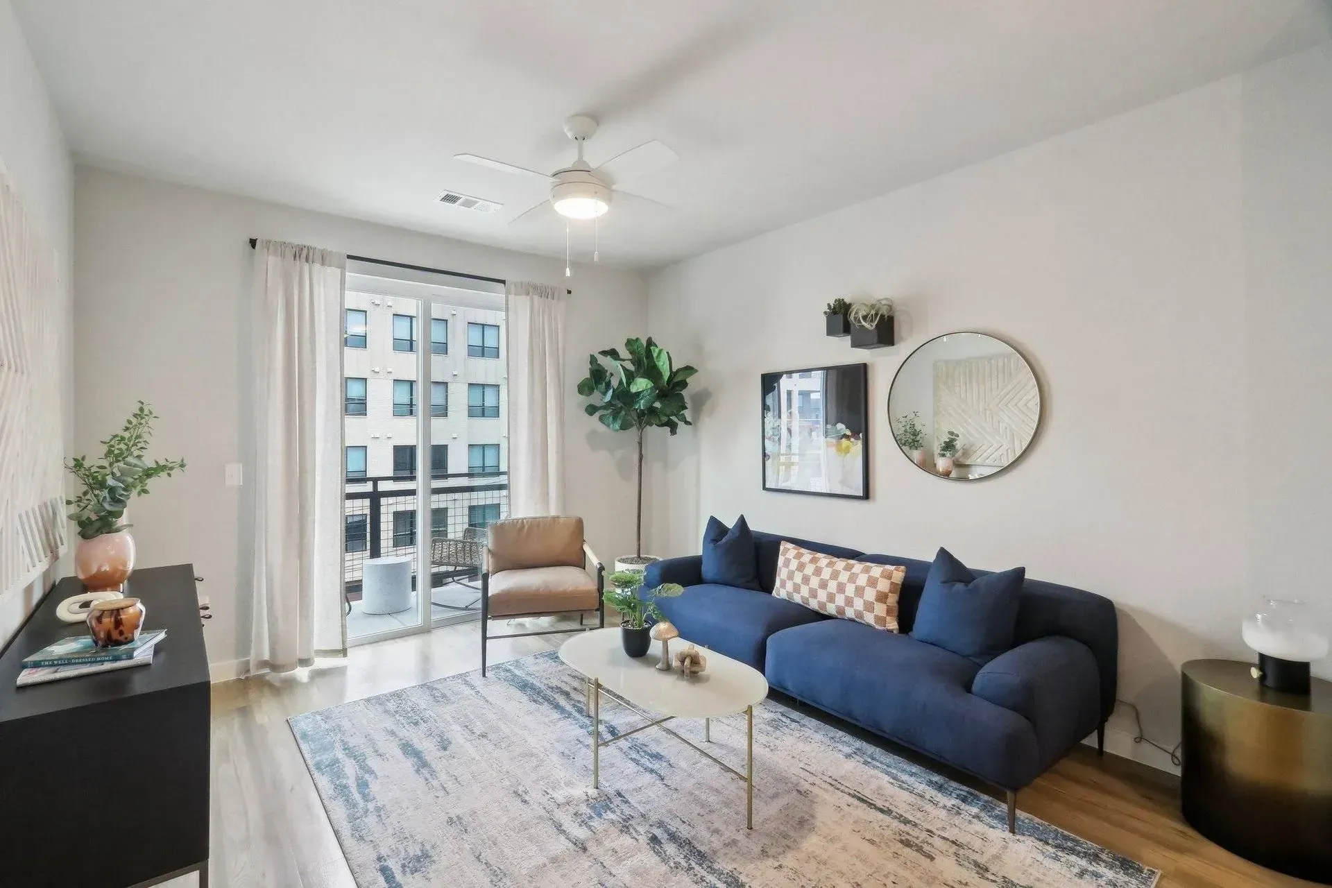 Living room with blue sofa, rug, and balcony access. White walls, wood floor, and gold accents.