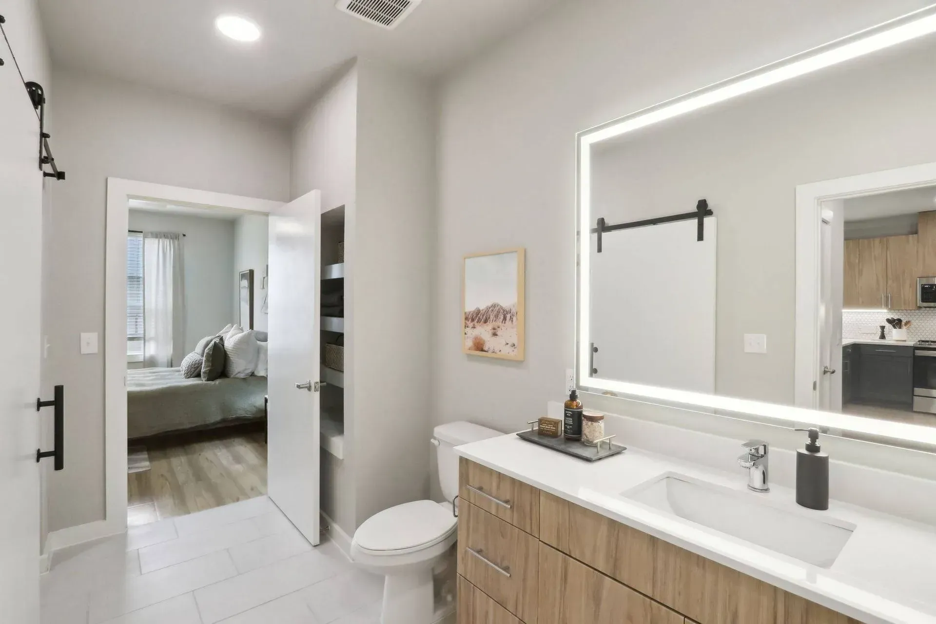 Bathroom with white tile, wood vanity, lit mirror, and doorway to bedroom.