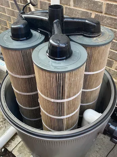 Three cylindrical pool filters inside a gray canister, with black caps on top, set outside against a brick wall.