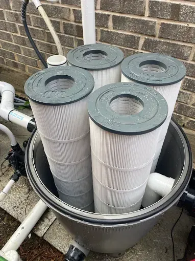 Four cylindrical pool filters inside a round filter housing, surrounded by plumbing against a brick wall.