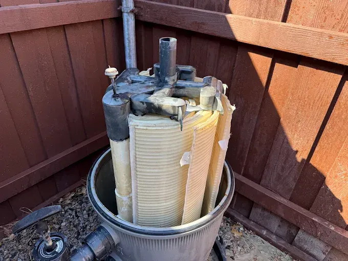 A cylindrical pool filter with a white pleated filter cartridge. The top of the filter is black and damaged. It's next to a wooden fence outdoors.