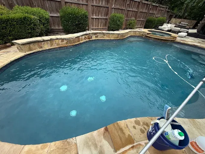 Swimming pool with blue water and stone coping; cleaning supplies on the deck, hot tub in the background.