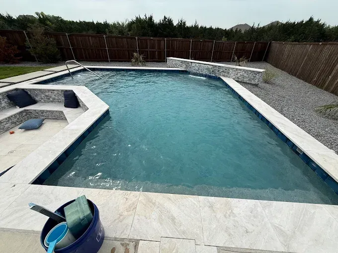 Swimming pool with blue water, white stone coping, and a stone waterfall feature. Pool cleaning supplies sit nearby.