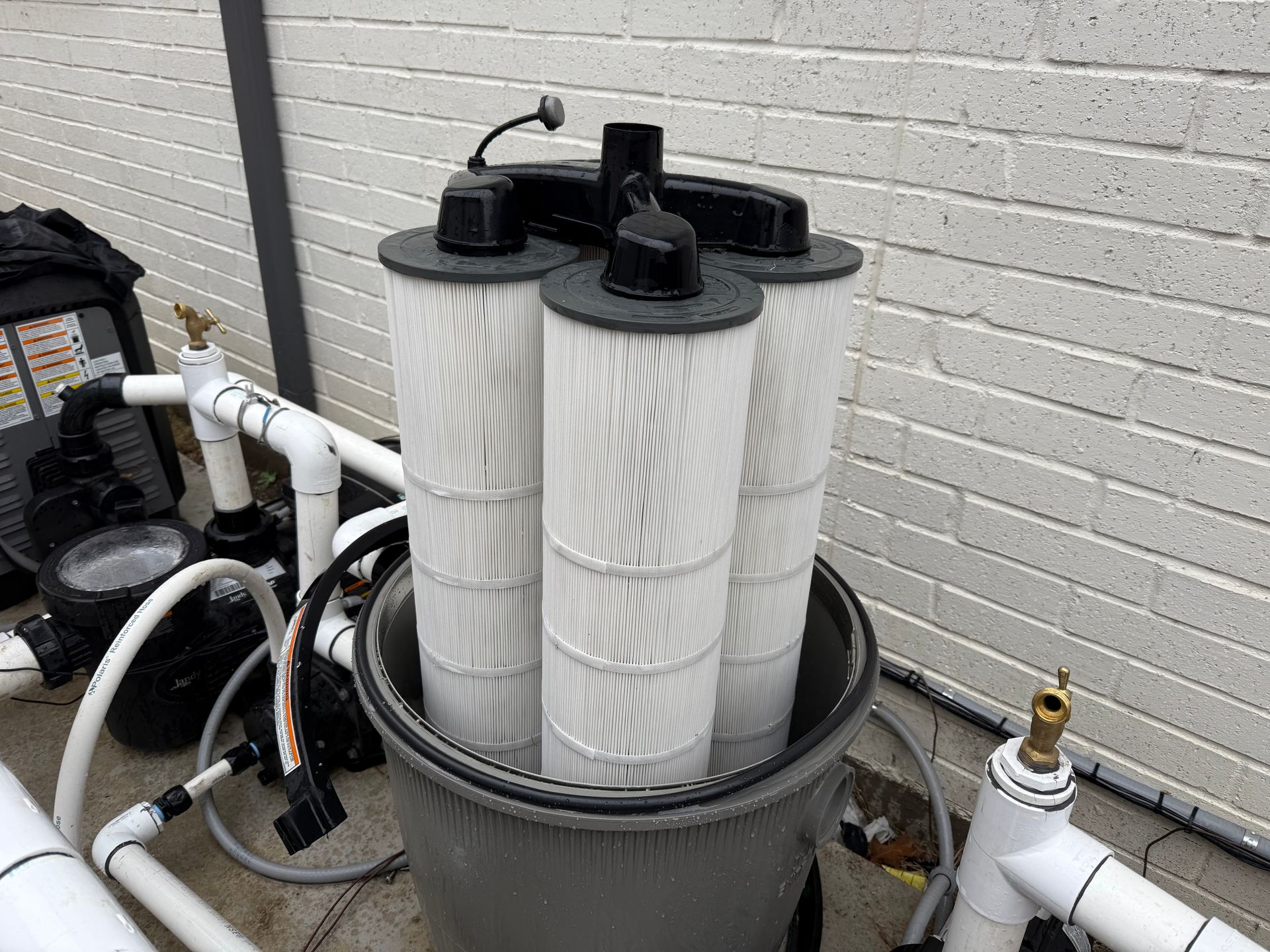 Three cylindrical pool filters in a gray housing, connected by white pipes and a black top. Set outdoors near a brick wall.