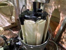 Pool filter cartridge removed from its housing for cleaning, outdoors. Cream-colored pleated filter, black top assembly, and gray housing.