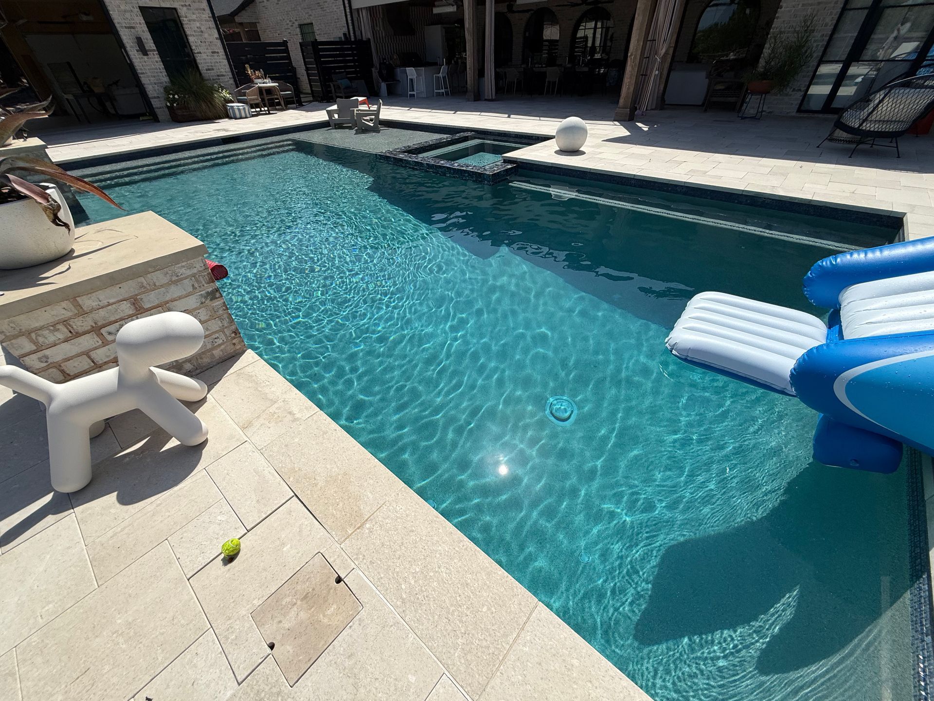 A rectangular swimming pool with clear blue water. A white dog statue sits on the edge, with a blue and white inflatable slide.