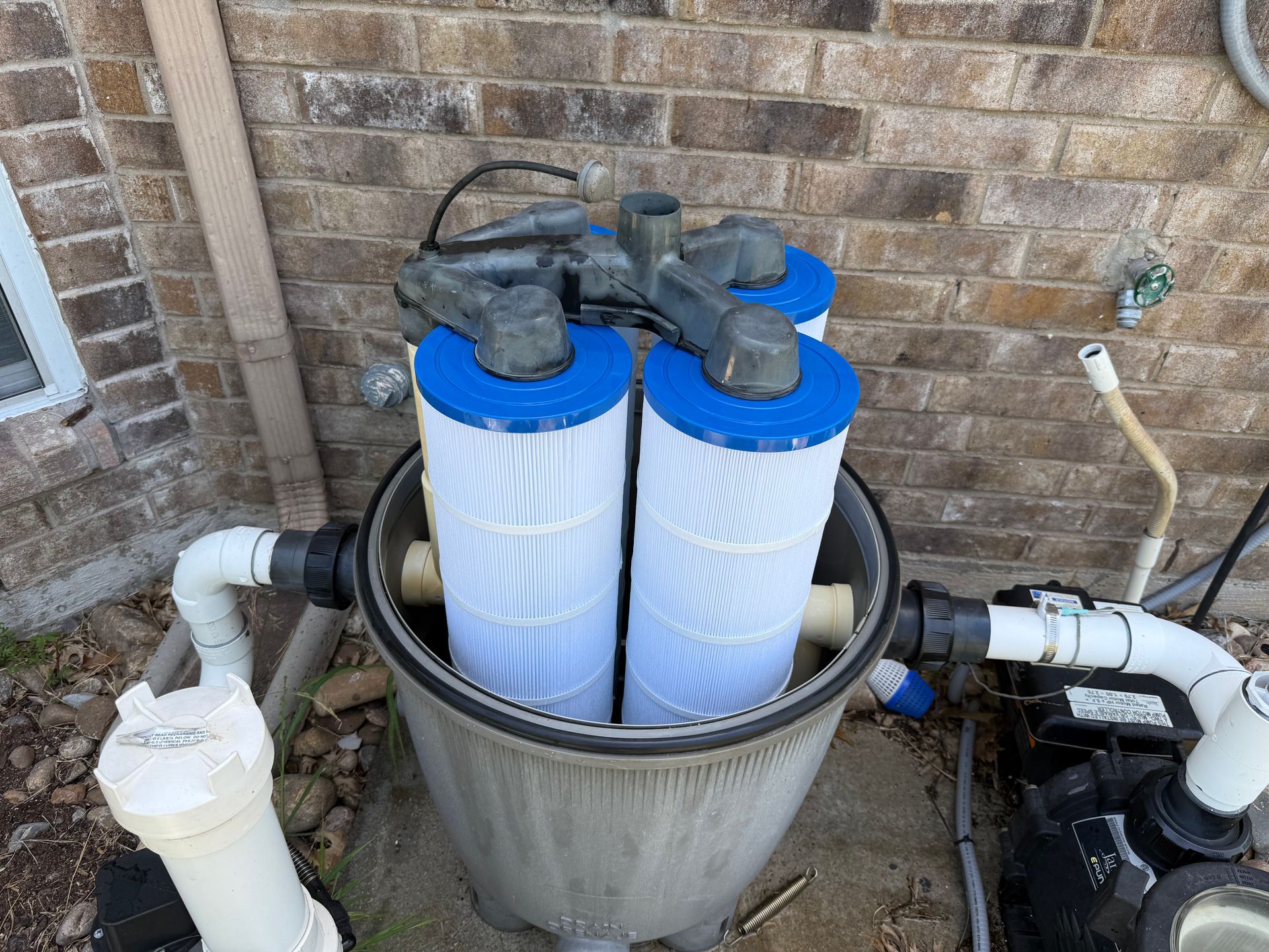 Pool filter system with two cylindrical filters inside a gray housing, near a brick wall. Blue caps top the filters, white pipes connect to the system.