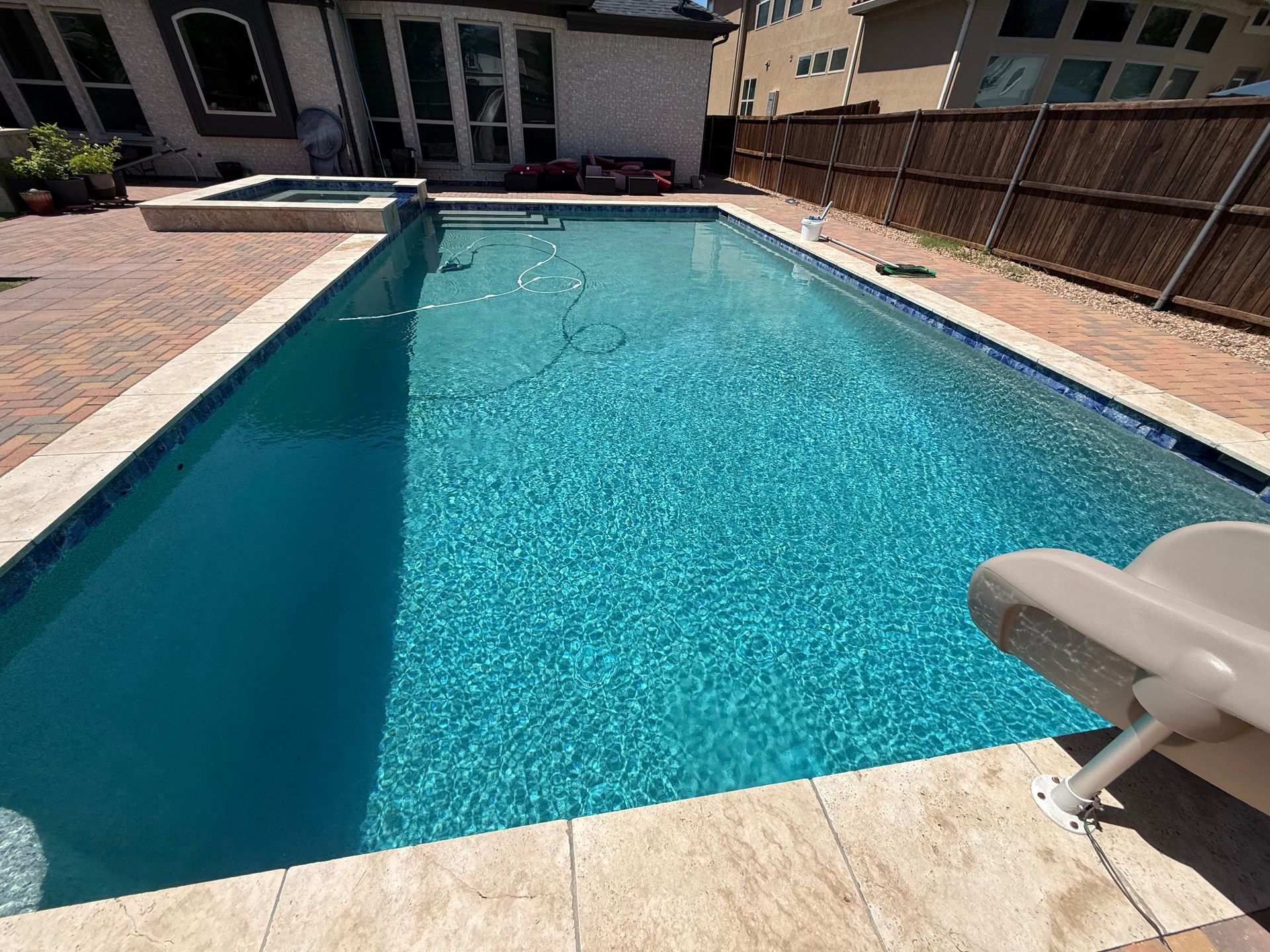 Swimming pool with clear blue water surrounded by beige tile. A small slide is visible on one side, and a spa is in the background.