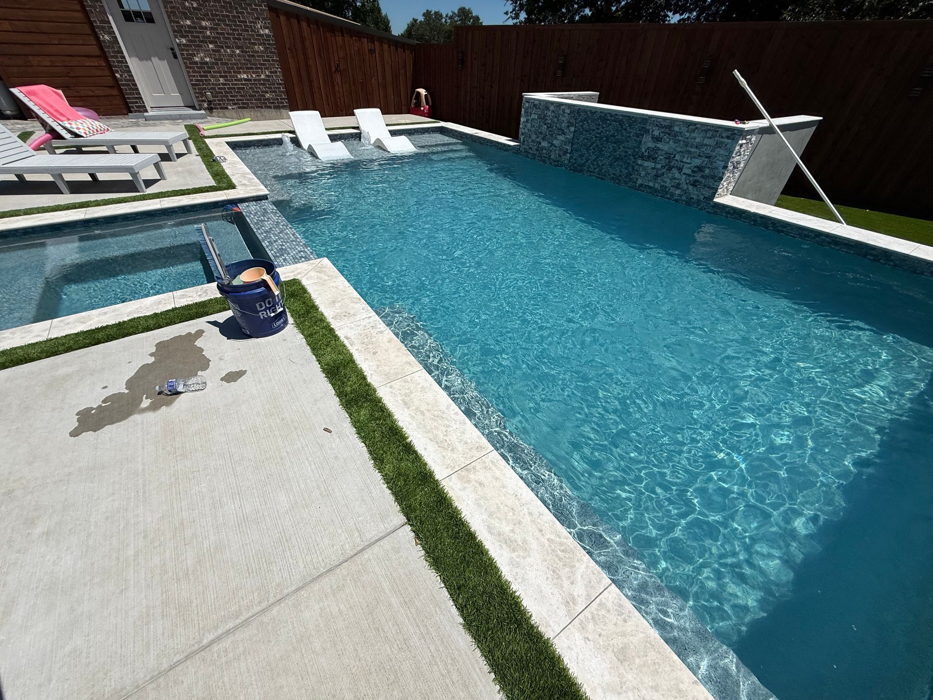 Modern rectangular pool with jacuzzi, lounge chairs, and water feature against a wooden fence, blue water, and light concrete.