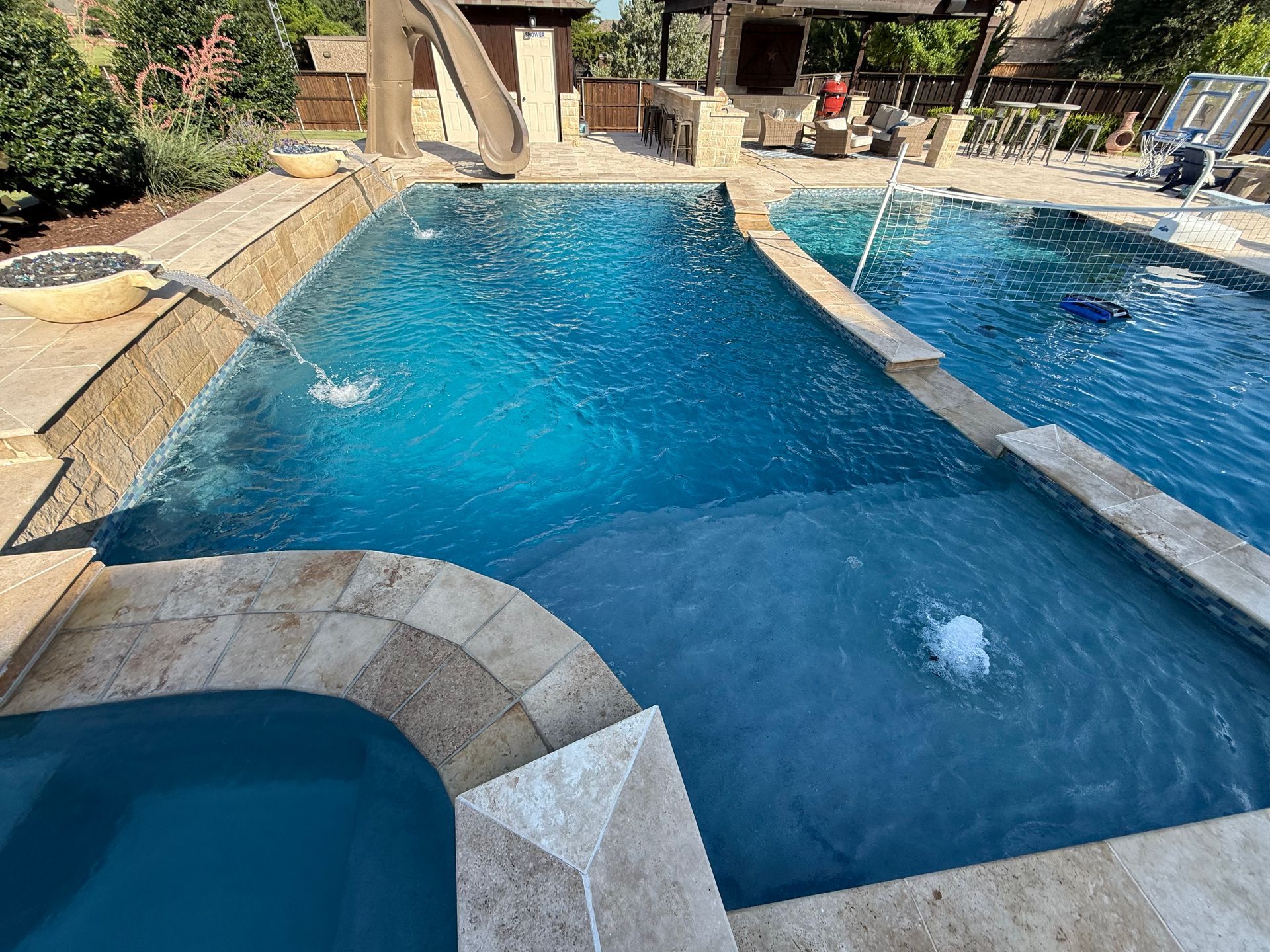 A backyard swimming pool with a slide and spa. Turquoise water contrasts with the light stone border and surrounding landscaping.