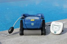 A dark blue robotic pool cleaner with a power supply sitting next to a swimming pool.
