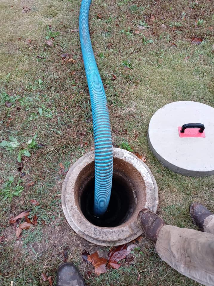 A septic tank being pumped. A blue hose enters the open tank, lid is nearby, and a person's feet are in frame.