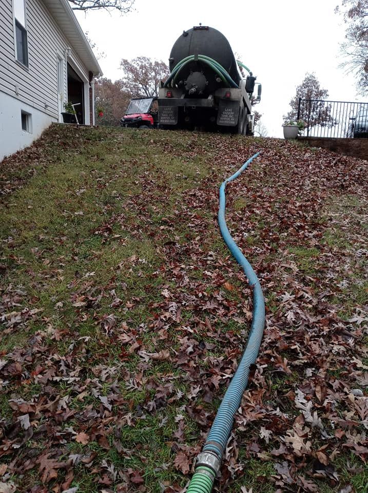 Septic tank truck with hose on a sloped, leaf-covered lawn next to a house.