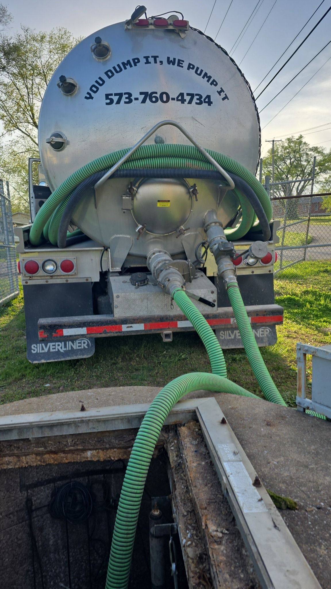 A septic truck with green hoses is parked over an open manhole.