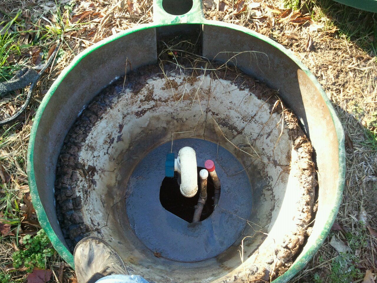 Open well with plumbing visible inside; green casing, dirt, and grass surround.