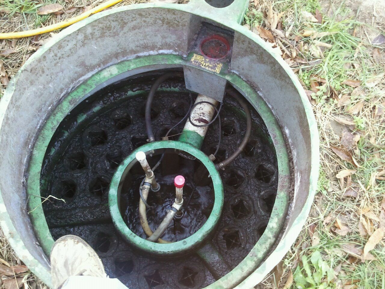 Open septic tank with green components, black interior, and a person's foot visible.