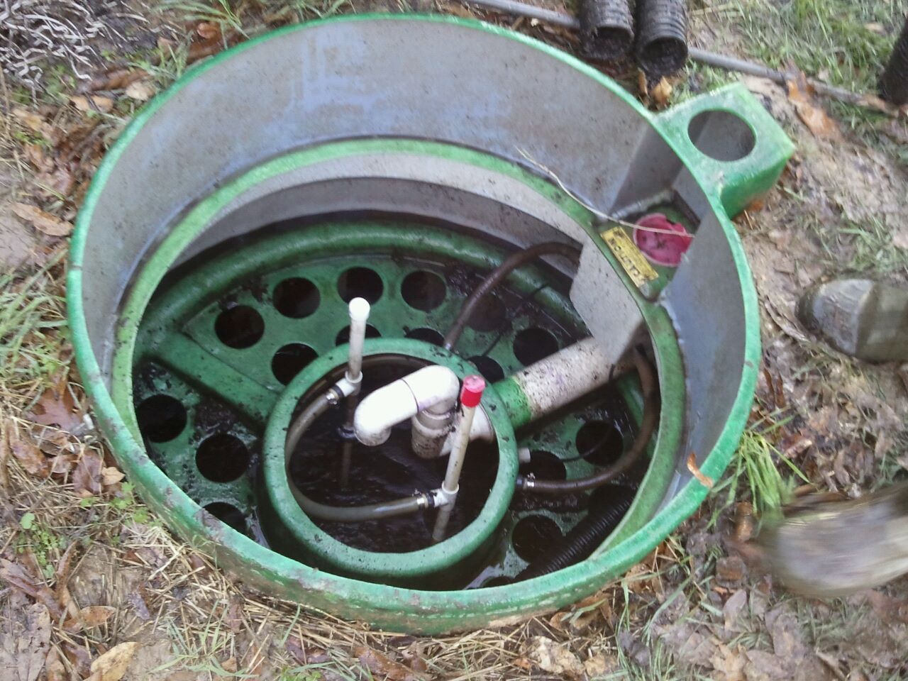 Open septic tank with green plastic components, pipes, and a white valve, set outdoors.