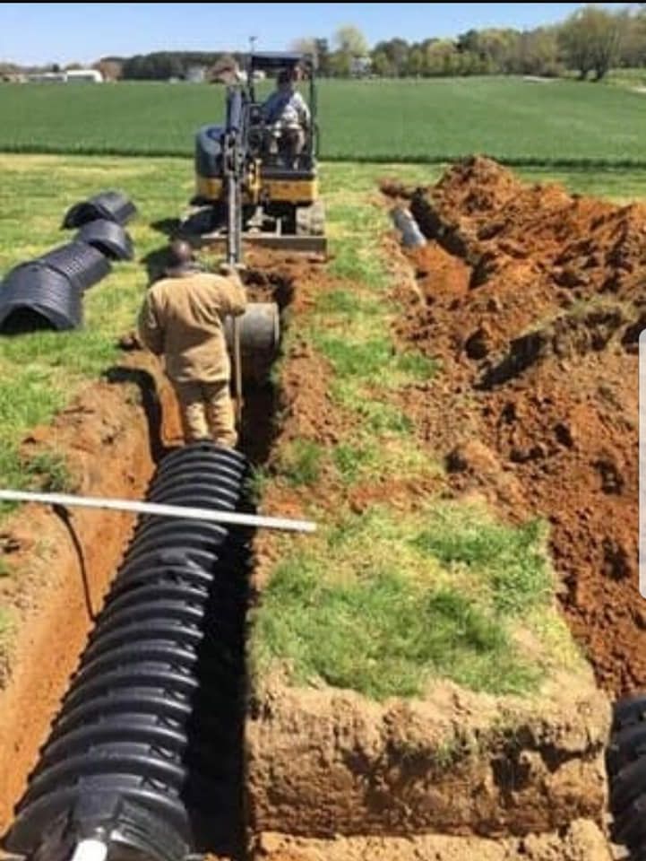 Workers installing corrugated black pipes in a trench using an excavator. Field setting.