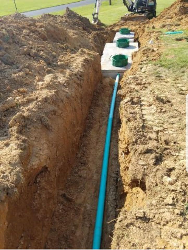 Trench with blue pipe leading to concrete septic tanks with green lids, in a grassy area.