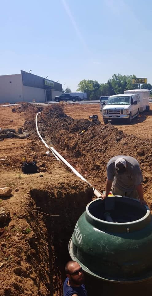 Construction workers installing septic tank in a dirt trench with white pipe and a white van parked nearby on a sunny day.