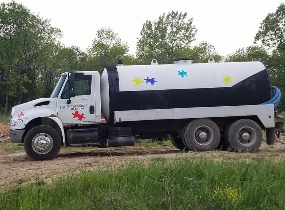 White septic truck with a black and white tank, adorned with colorful star shapes, parked on a dirt road.