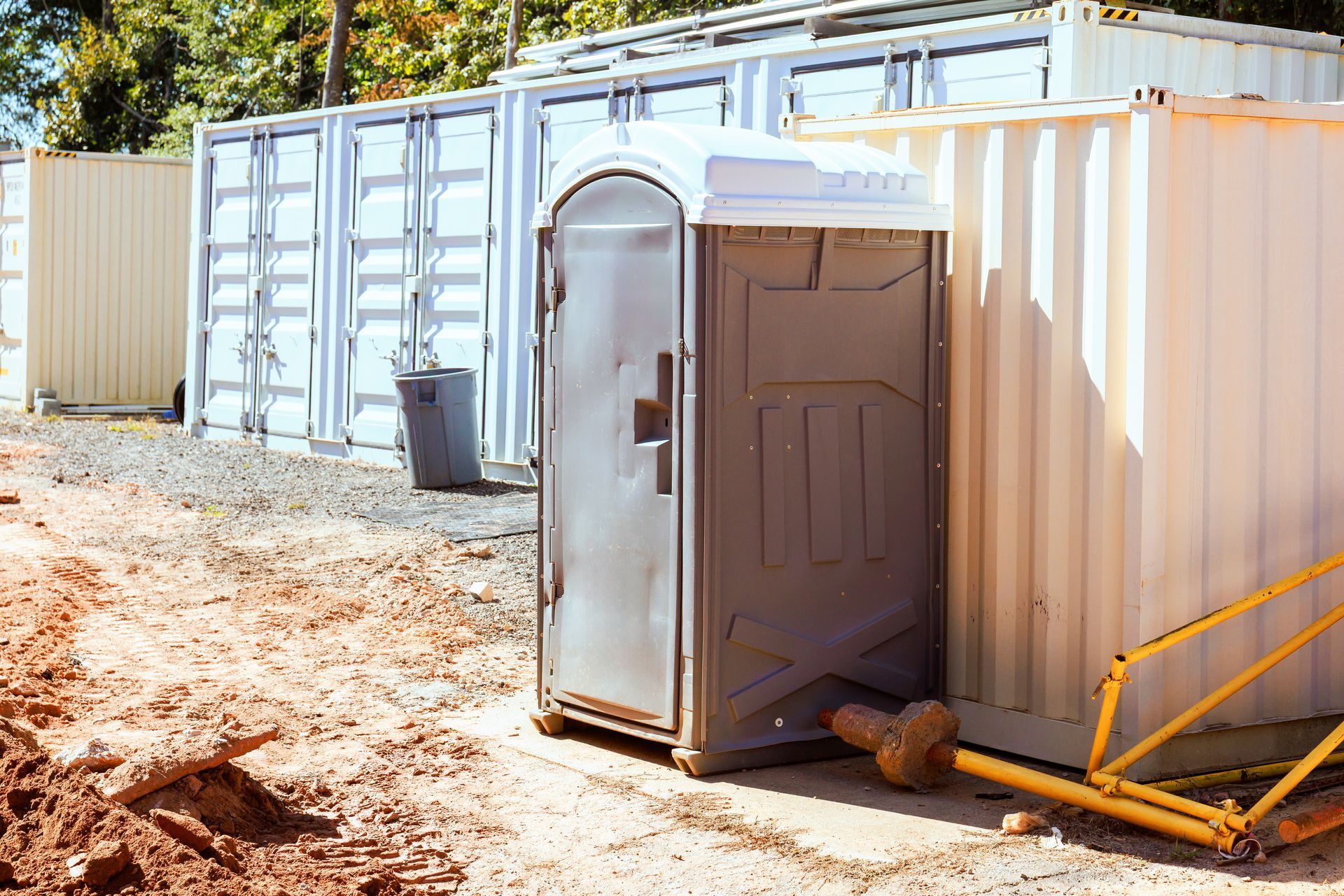 Gray portable toilet in front of several white metal storage containers on a construction site.