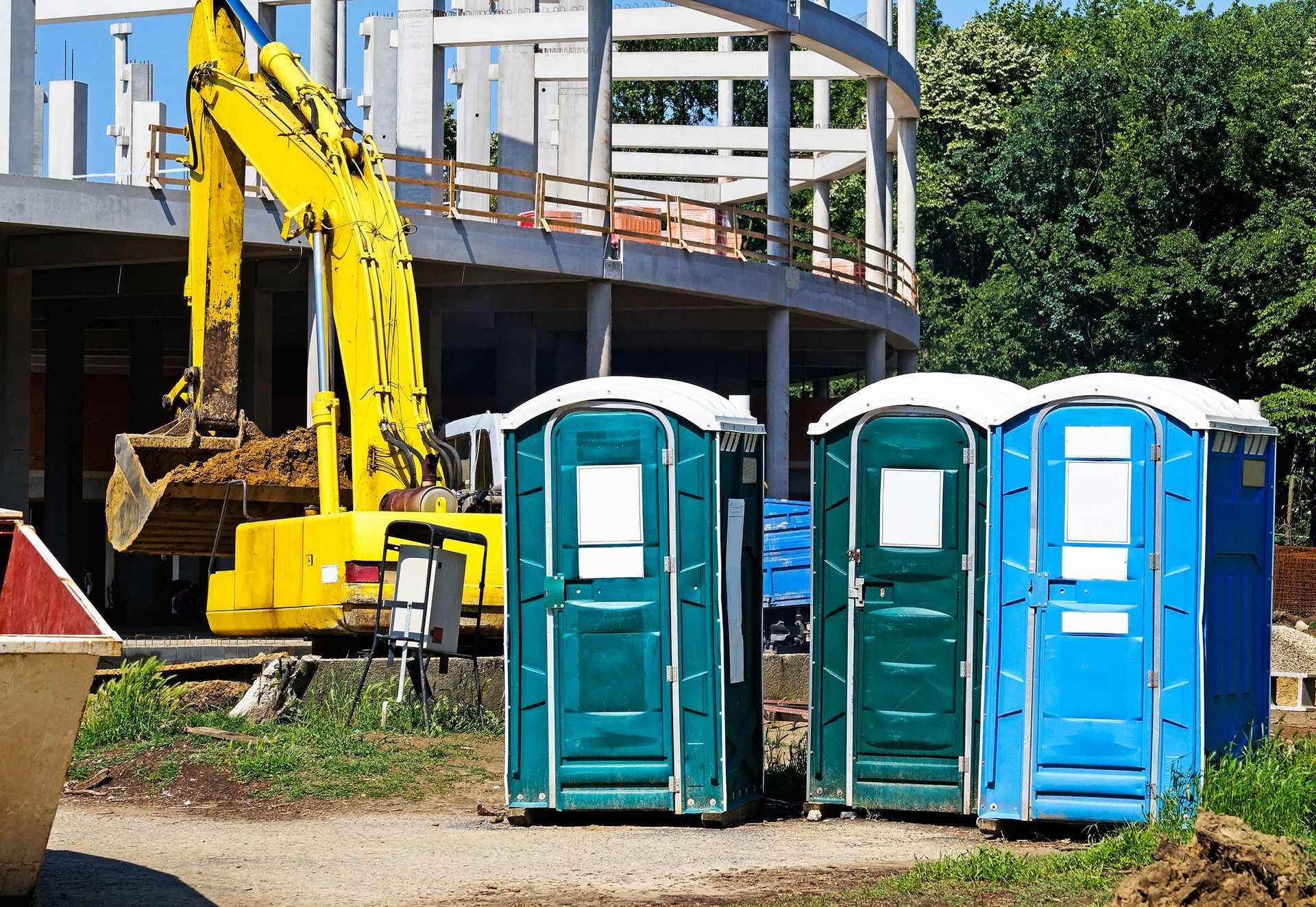 Three portable toilets in front of a building under construction; a yellow excavator operates in the background.