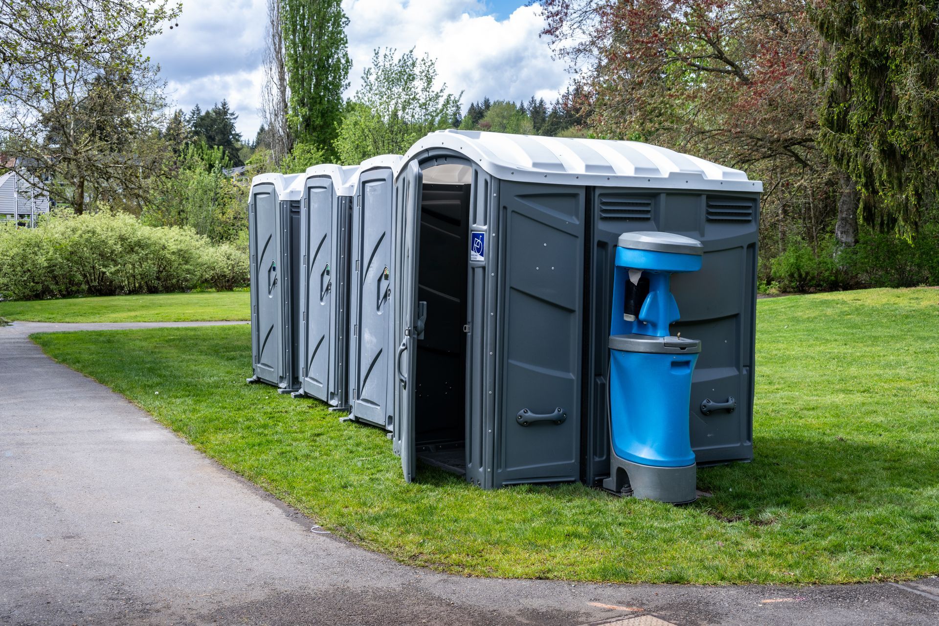 Portable toilets and handwashing station on grass next to a paved path in a park.